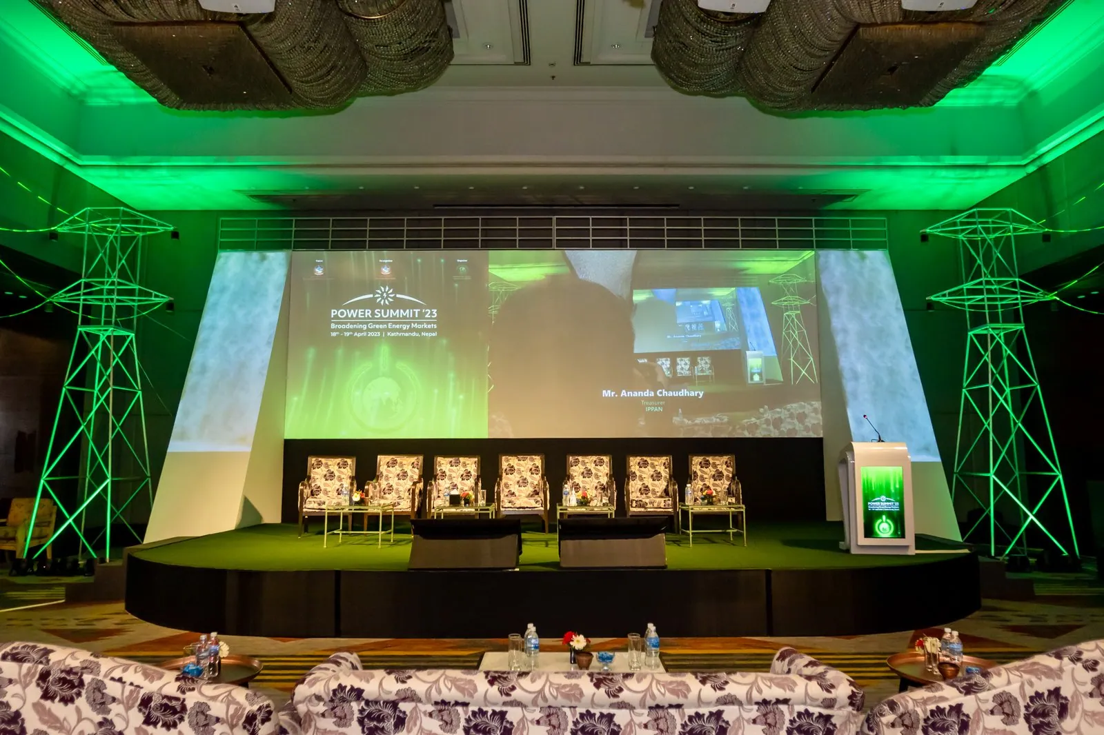 The empty Power Summit '23 stage: two green lit transmission-tower structures, a wide screen and angled side screens, and rows of chairs on green turf.