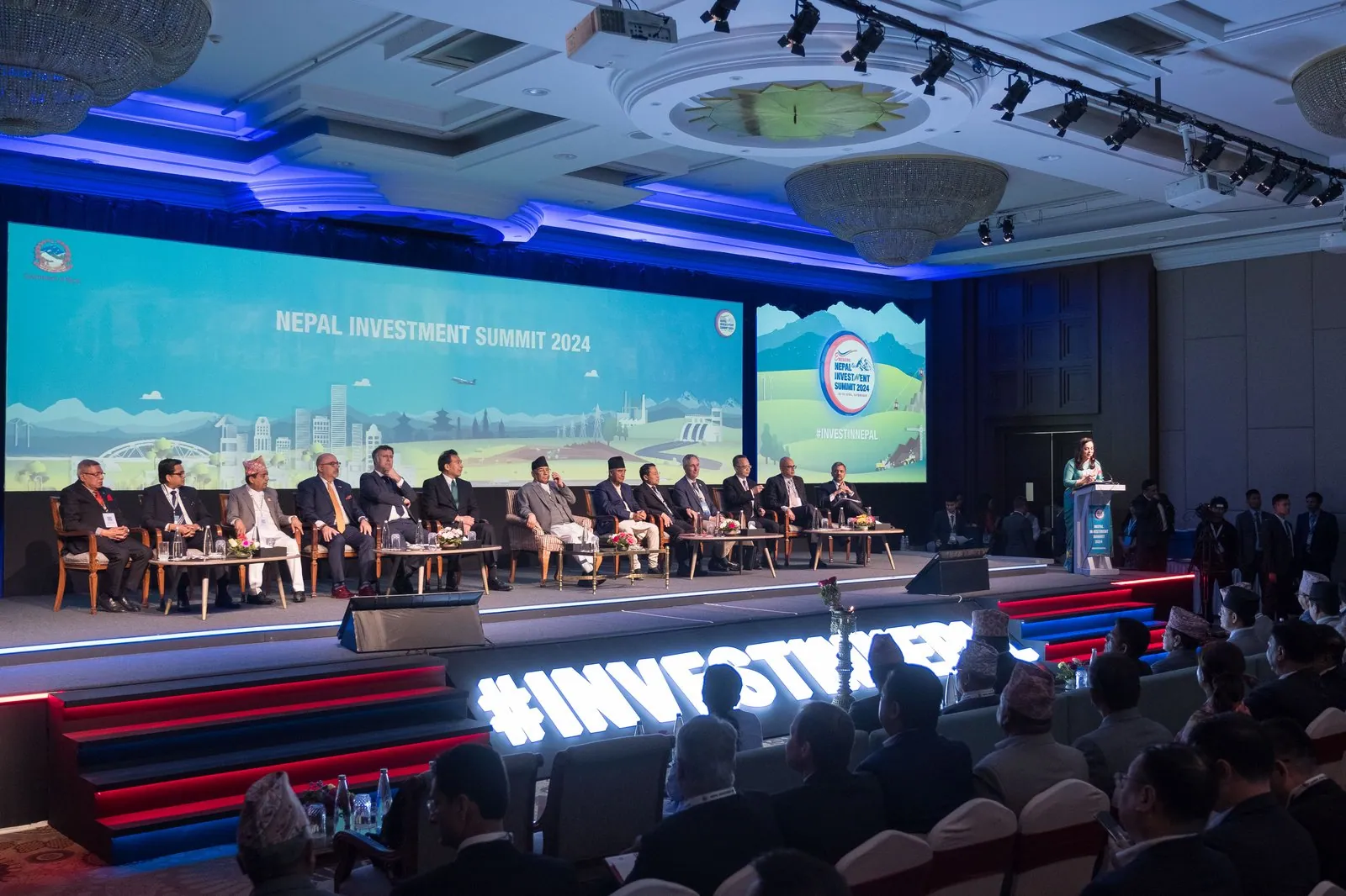 Nepal Investment Summit 2024 stage: a wide screen with the summit title over a landscape illustration, a row of guests seated on stage and the hashtag InvestInNepal in lit letters on the stage front.