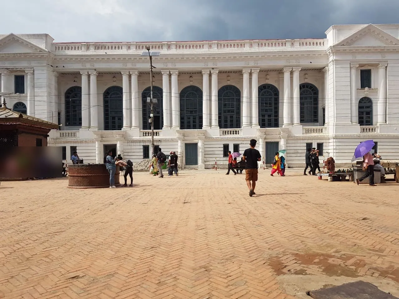 Daytime photograph of the white Gaddi Baithak facade across a brick-paved square: two pedimented end pavilions and a central run of seven arched windows with dark blue frames between paired columns.