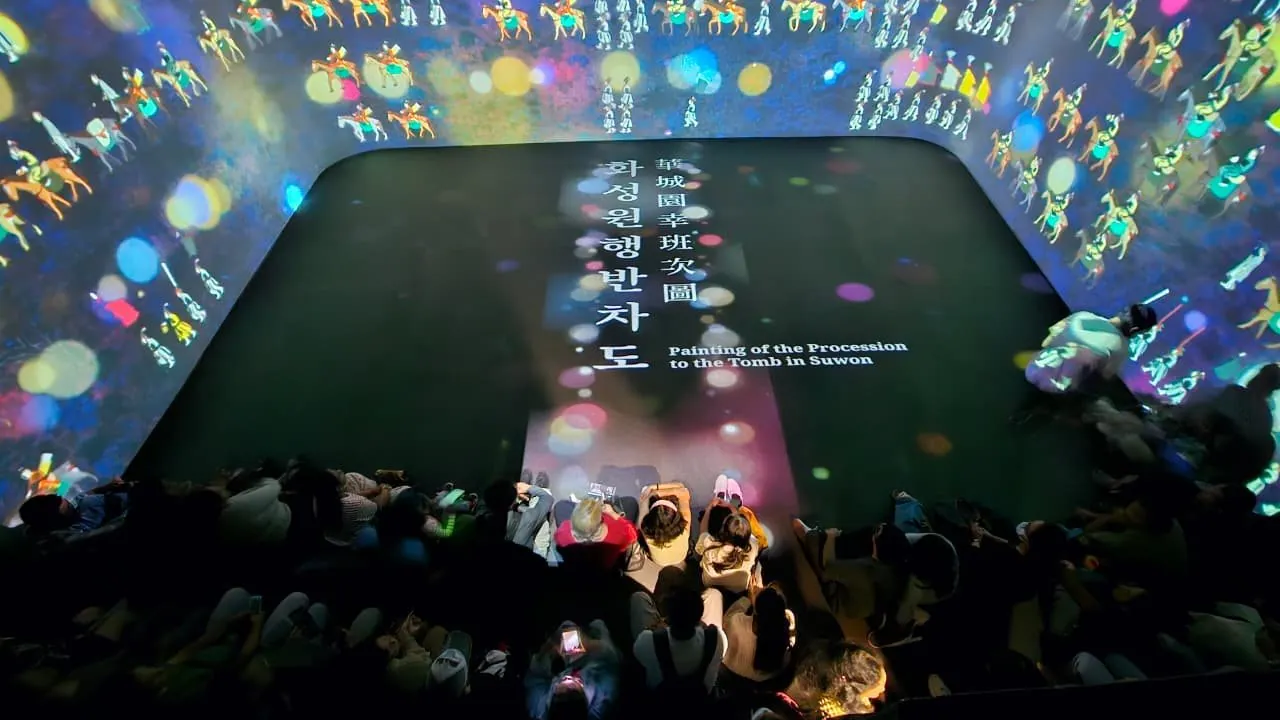 View from above of the projected room: the floor carries the title “Painting of the Procession to the Tomb in Suwon” in Korean and English, surrounded by walls of mounted riders and coloured light, with visitors seated at the bottom of the frame.