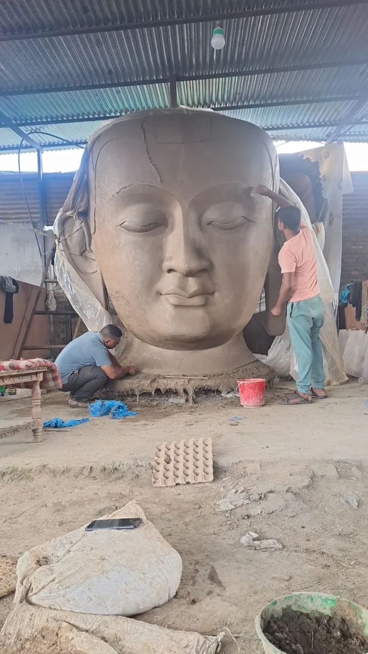 A large clay Buddha head in a corrugated-roof workshop, with two people working on it.