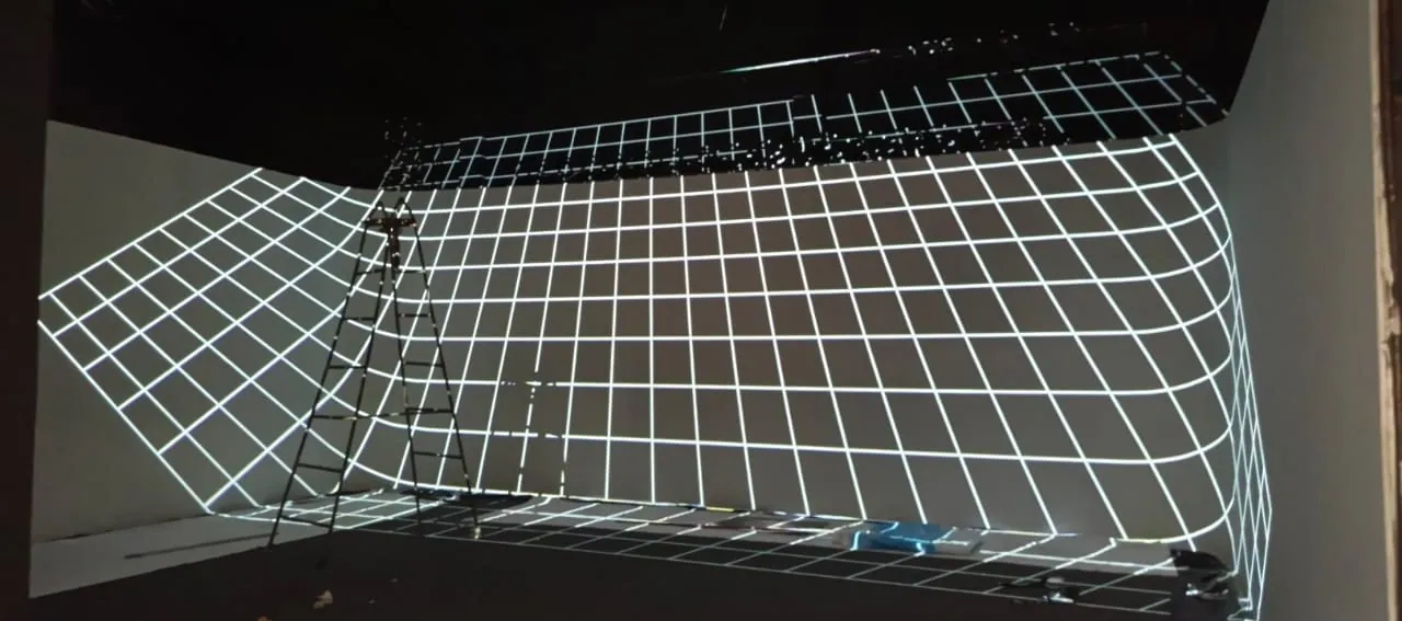 A white grid projected across two walls and the floor, bending around the rounded corner, with a ladder standing in front.