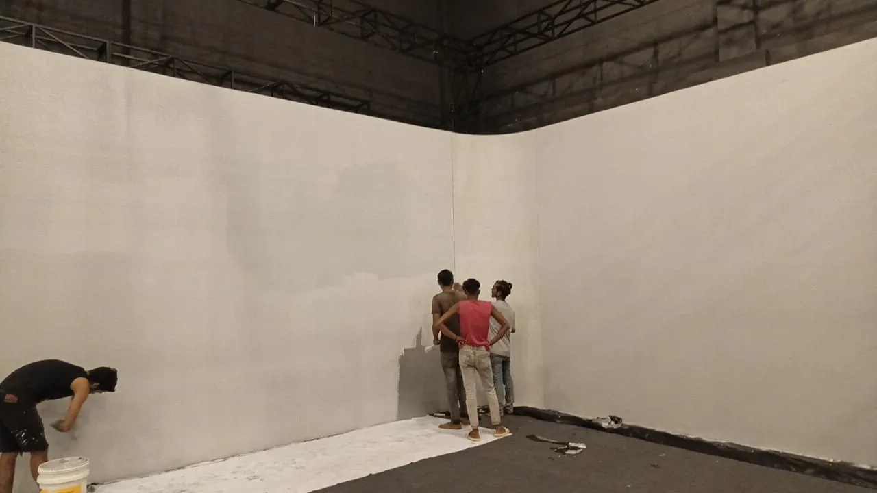 Workers painting the tall ply-board screen walls of the hall, with a rounded corner between two walls.