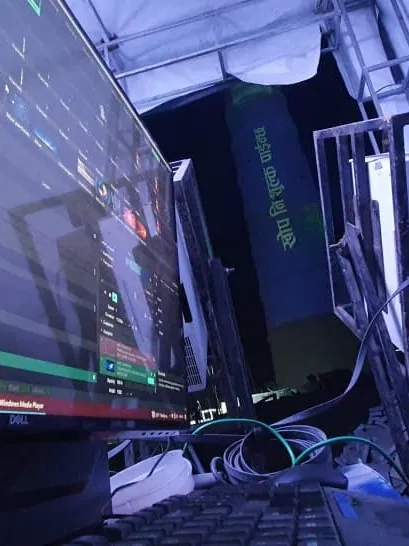A widescreen monitor showing a clip timeline and a projector control window, with a projector on a steel rack and the tower in the background.