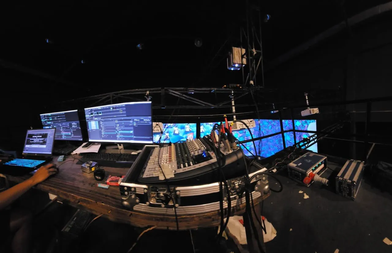 Wide view of the control desk: two monitors running playout software, a laptop and an audio mixer, with the projected screens behind.